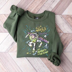 May include: An olive green sweatshirt featuring a graphic of Buzz Lightyear from Toy Story. The design includes the text "Buzz Lightyear" and "To Infinity and Beyond" with a space-themed background. The sweatshirt has long sleeves.