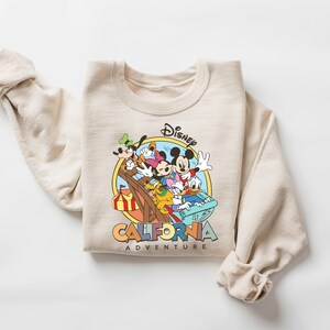 May include: Beige crewneck sweatshirt featuring a colorful graphic of Mickey Mouse, Minnie Mouse, Donald Duck, Goofy, and Pluto. The graphic is set within a yellow circle and includes the text "Disney California Adventure".