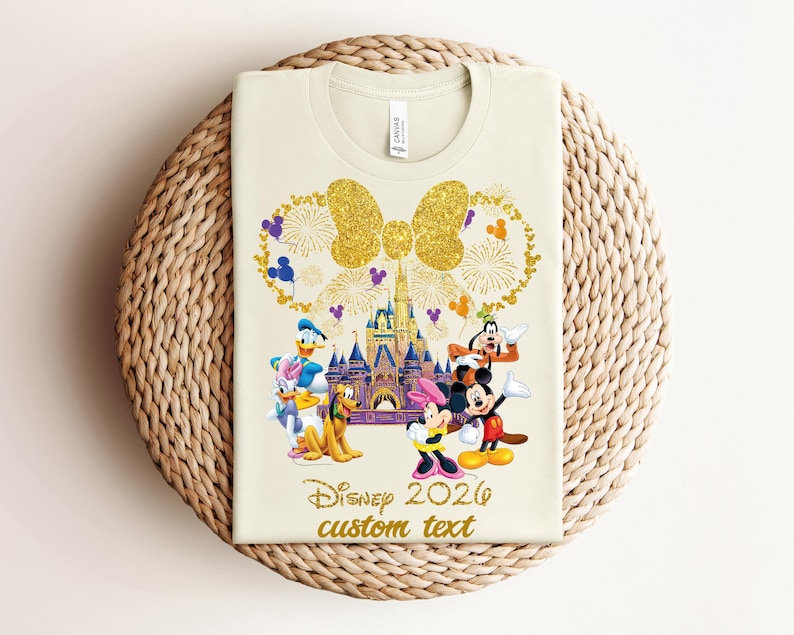 K&ouml;nnte beinhalten: Cremefarbenes T-Shirt mit goldener Glitzer-Schleife und Feuerwerksdesign. Das Shirt zeigt Disney-Figuren wie Mickey und Minnie Mouse, Donald Duck und Goofy, mit dem Text "Disney 2026 custom text".