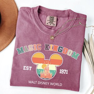 May include: A maroon t-shirt with a crew neck featuring a colorful graphic of Mickey Mouse ears with a sunset design and a castle. The text "MAGIC KINGDOM EST 1971 WALT DISNEY WORLD" is printed below the graphic. A gold necklace is draped across the shirt.