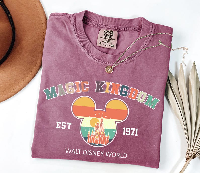May include: A maroon t-shirt with a crew neck featuring a colorful graphic of Mickey Mouse ears with a sunset design and a castle. The text "MAGIC KINGDOM EST 1971 WALT DISNEY WORLD" is printed below the graphic. A gold necklace is draped across the shirt.