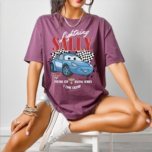 May include: A mauve-colored t-shirt featuring a graphic of a blue cartoon car with the text "Lightning Sally" above it. The design includes checkered flags and the words "Piston Cup Racing Series 3 Time Champ".