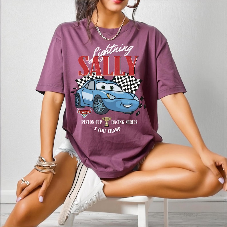May include: A mauve-colored t-shirt featuring a graphic of a blue cartoon car with the text "Lightning Sally" above it. The design includes checkered flags and the words "Piston Cup Racing Series 3 Time Champ".