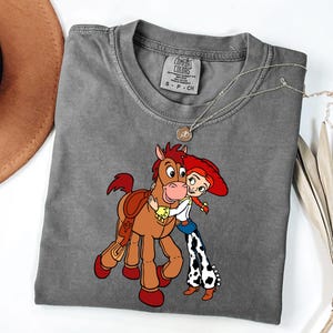 May include: A grey Comfort Colors t-shirt with a cartoon illustration of a brown horse embracing a cowgirl with red hair. The cowgirl wears a white shirt, blue jeans, and black and white patterned trousers. A silver necklace is draped across the shirt.