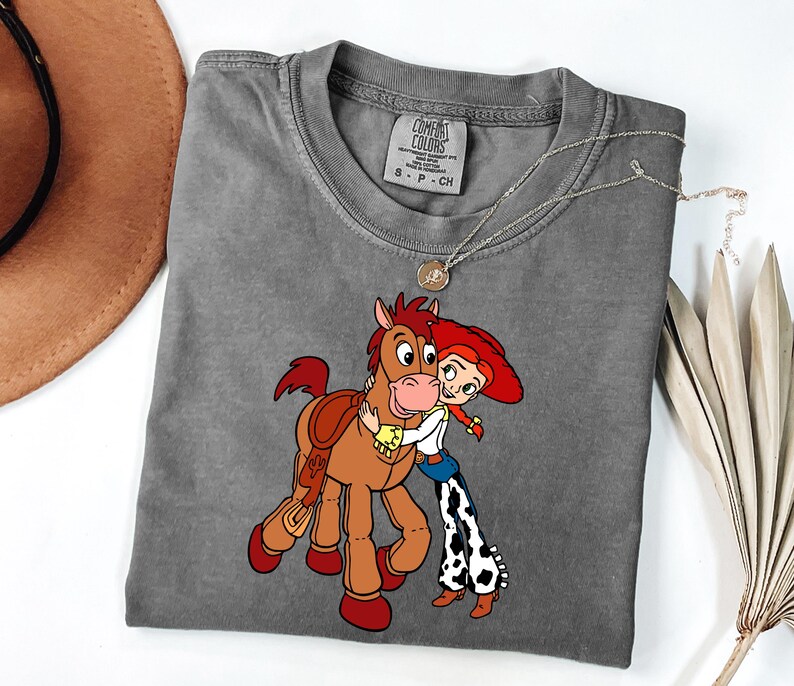 May include: A grey Comfort Colors t-shirt with a cartoon illustration of a brown horse embracing a cowgirl with red hair. The cowgirl wears a white shirt, blue jeans, and black and white patterned trousers. A silver necklace is draped across the shirt.