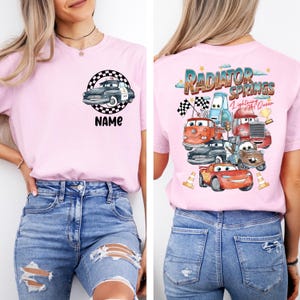 May include: A light pink t-shirt featuring a cartoon police car design inside a checkered circle with the word "NAME" below it. The back of the shirt displays a colorful graphic of characters from the movie "Cars" with the text "Radiator Springs".