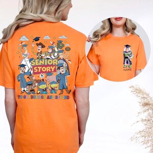 May include: Orange t-shirt with a graduation design featuring animated film characters. The back of the shirt has the text "SENIOR STORY" and "TO GRADUATION AND BEYOND". A smaller image of Buzz Lightyear is on the front.