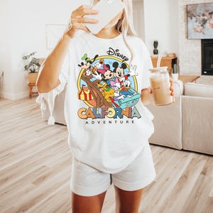 May include: White t-shirt featuring a colorful graphic of Disney characters, including Mickey Mouse, Minnie Mouse, and Donald Duck, with the text "California Adventure". The design is set within a circular frame.