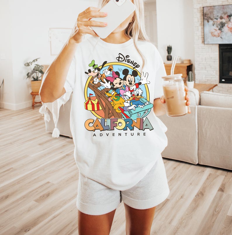 May include: White t-shirt featuring a colorful graphic of Disney characters, including Mickey Mouse, Minnie Mouse, and Donald Duck, with the text "California Adventure". The design is set within a circular frame.
