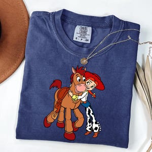 May include: A navy blue Comfort Colors t-shirt with a cartoon illustration of a horse and a cowgirl embracing. The shirt has a crew neck and short sleeves. A gold necklace with a pendant is draped on the shirt.