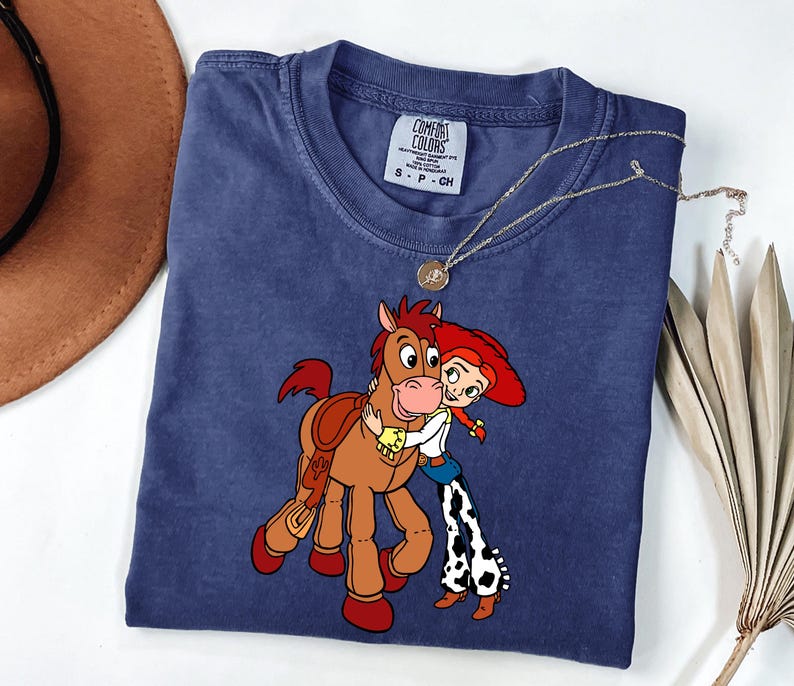 May include: A navy blue Comfort Colors t-shirt with a cartoon illustration of a horse and a cowgirl embracing. The shirt has a crew neck and short sleeves. A gold necklace with a pendant is draped on the shirt.