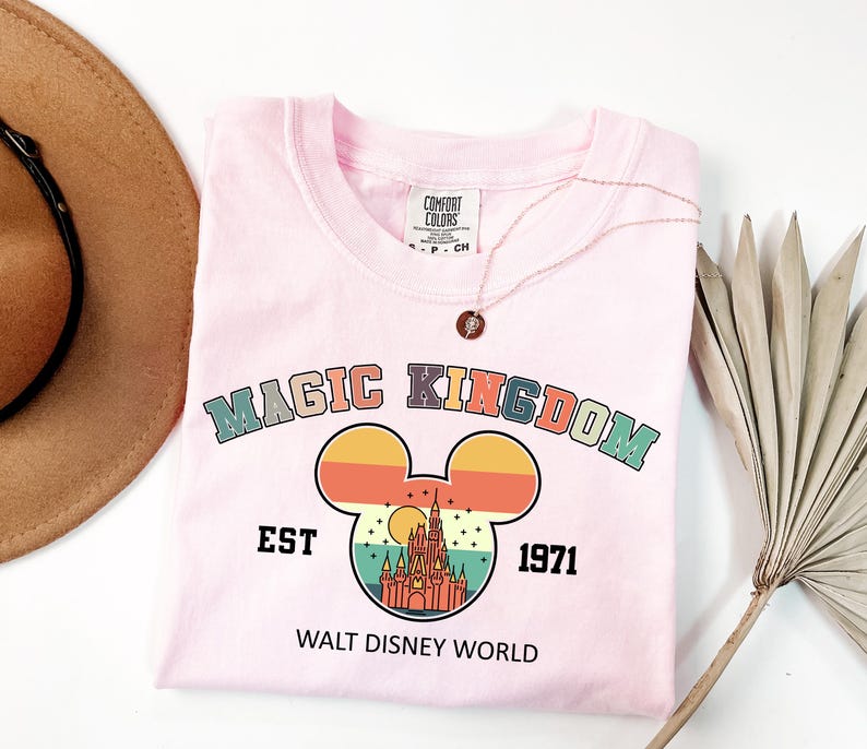 May include: A light pink t-shirt with the words "MAGIC KINGDOM" in colorful letters above a Mickey Mouse head design. The design includes a castle and the text "EST 1971 WALT DISNEY WORLD". A gold necklace is on the shirt.