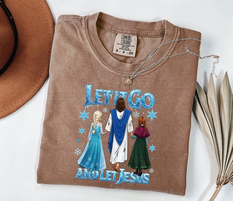 May include: Brown t-shirt with the text "LET IT GO AND LET JESUS" and a graphic of Jesus with two cartoon characters. The shirt has a crew neck and short sleeves. A silver necklace with a pendant is on the shirt.