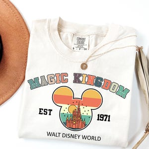 May include: Cream-colored t-shirt with a retro design featuring the words "MAGIC KINGDOM" above a Mickey Mouse silhouette. The silhouette contains a castle illustration, the text "EST 1971", and "WALT DISNEY WORLD". A gold necklace rests on the shirt.