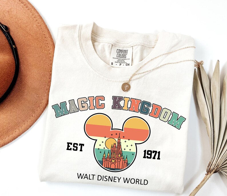 May include: Cream-colored t-shirt with a retro design featuring the words "MAGIC KINGDOM" above a Mickey Mouse silhouette. The silhouette contains a castle illustration, the text "EST 1971", and "WALT DISNEY WORLD". A gold necklace rests on the shirt.