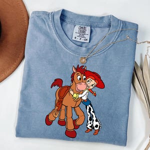 May include: A light blue Comfort Colors t-shirt with a cartoon graphic of a horse and a cowgirl embracing. The shirt has a round neckline and short sleeves. A gold necklace is draped across the shirt.