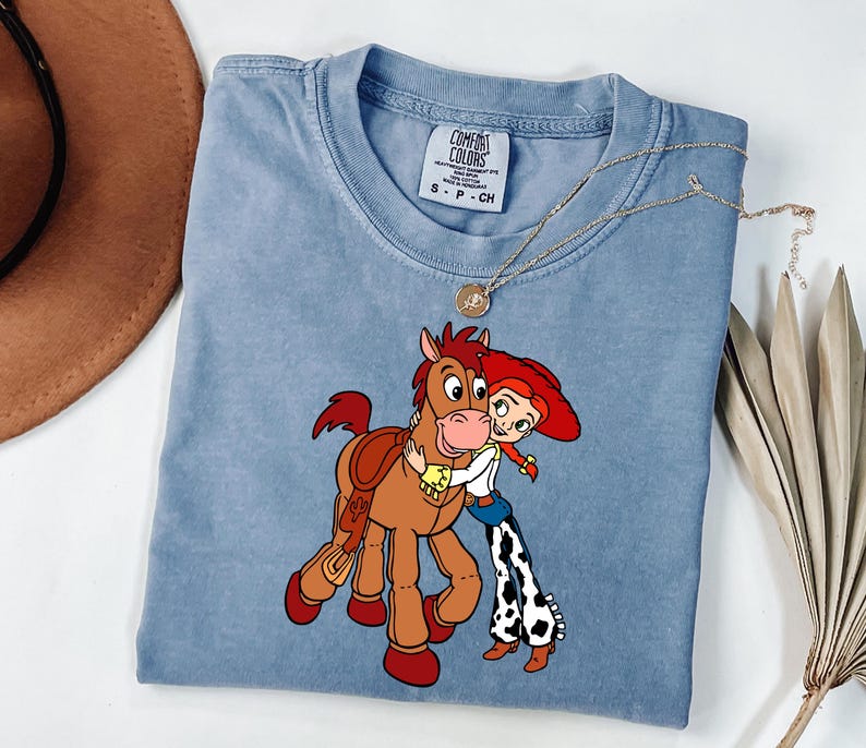 May include: A light blue Comfort Colors t-shirt with a cartoon graphic of a horse and a cowgirl embracing. The shirt has a round neckline and short sleeves. A gold necklace is draped across the shirt.