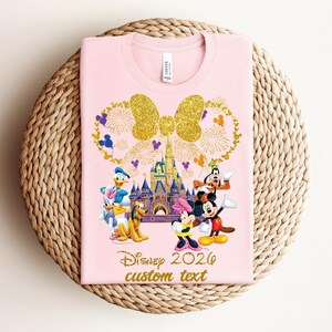 K&ouml;nnte beinhalten: Hellrosa T-Shirt mit goldener Glitzer-Schleife und Feuerwerk-Design, Disney-Figuren und einem Schloss. Der Text "Disney 2020 custom text" ist in Gold gedruckt.