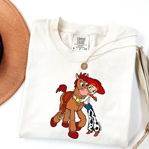 May include: Cream-coloured t-shirt featuring a cartoon illustration of a character from Toy Story hugging a horse. The character wears a red hat, white shirt, and blue jeans with black and white cow print chaps. A gold necklace is draped on the shirt.