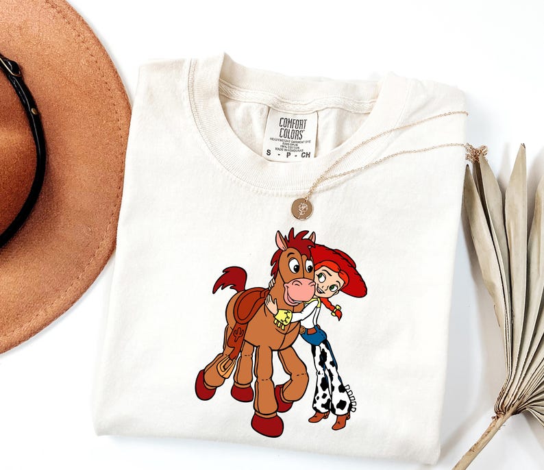 May include: Cream-coloured t-shirt featuring a cartoon illustration of a character from Toy Story hugging a horse. The character wears a red hat, white shirt, and blue jeans with black and white cow print chaps. A gold necklace is draped on the shirt.