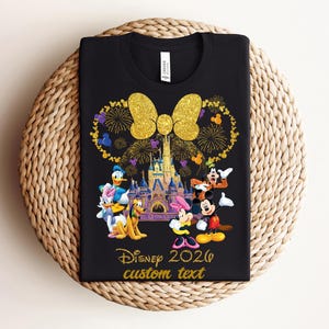 K&ouml;nnte beinhalten: Schwarzes T-Shirt mit einem Disney-Design mit Micky und Minnie Maus, Donald Duck und Goofy vor einem Schloss. Das Design beinhaltet eine goldene Schleife, Feuerwerk und den Text "Disney 2026 custom text."