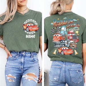 May include: Green t-shirt featuring a cartoon car design with the text "NAME" on the front. The back of the shirt displays a graphic of various cartoon cars with the text "RADIATOR SPRINGS Lightning McQueen".