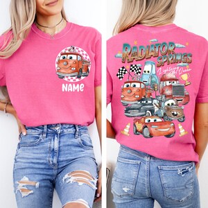 May include: Pink t-shirt featuring a cartoon fire truck design on the front and a Radiator Springs graphic on the back. The front design includes the word "NAME" below the fire truck. The back design features various car characters.