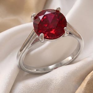 May include: A silver ring featuring a large, round, faceted deep red gemstone held in a four-prong setting. The band is smooth and polished, showcasing a clean, metallic finish that contrasts with the vibrant, saturated color of the central stone.