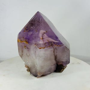 May include: A large, raw amethyst crystal with a pointed top. The crystal features a gradient of colors, from deep purple at the top to a translucent, pale lavender at the base. Some areas have golden-brown inclusions.