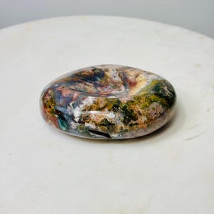 May include: A polished, oval-shaped stone with a natural, multicolored pattern. The stone features shades of brown, green, red, and white, creating a unique, organic appearance. The stone is smooth and appears to be a decorative item.