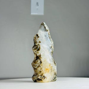 May include: A natural, polished stone sculpture with a unique, organic shape. The stone features a mix of translucent white, brown, and yellow hues, creating a striking visual contrast. The sculpture stands upright, showcasing its natural beauty.