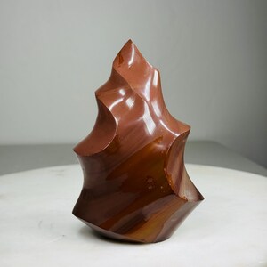 May include: A polished, abstract sculpture in shades of brown and reddish-brown. The art piece has a unique, organic shape with curved, flowing lines and a glossy finish. The sculpture is displayed on a white surface, with a neutral background.