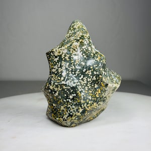 May include: A polished, freeform gemstone sculpture. The stone features a speckled pattern of dark green, white, and yellow. The sculpture has a rounded, organic shape with a pointed top and a textured surface. It is displayed on a white surface.