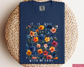 May include: Navy blue t-shirt featuring a floral design with orange, yellow, pink, and blue flowers. The text "IT IS WELL WITH MY SOUL" is printed in white. The shirt is made of a soft material.