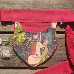 Puede incluir: Una envoltura de tela roja con un bolsillo que presenta una ilustración colorida de una sirena y un unicornio. La ilustración incluye pájaros, flores y peces.