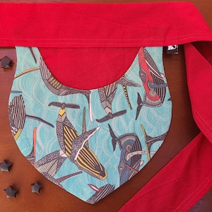 May include: A turquoise blue fabric bib with a red trim and a pattern of black and white stylized fish. The bib is tied with a red fabric strap.
