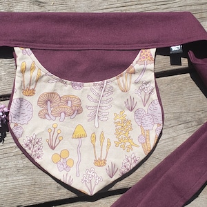 May include: A burgundy head wrap with a beige panel featuring a mushroom and fern pattern in shades of orange, yellow, and purple. The wrap is lying on a wooden surface, with a sprig of purple flowers.