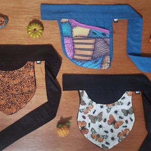 Può includere: Quattro fasce per capelli in tessuto con diversi motivi. La prima fascia è blu con una tasca a patchwork. La seconda fascia è nera con un motivo a zucca arancione. La terza fascia è nera con un motivo a farfalla bianca. La quarta fascia è nera con un motivo floreale bianco e arancione.