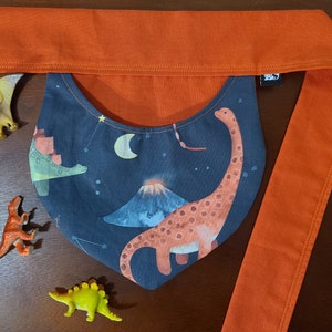 Puede incluir: Un babero de tela azul oscuro con un diseño de dinosaurio, volcán y estrellas. El babero está unido a una larga correa de tela naranja.