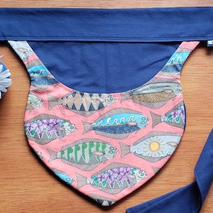 May include: A pink fabric baby carrier with a navy blue strap. The carrier has a pattern of colorful fish on a pink background. The strap is long and can be tied around the waist.