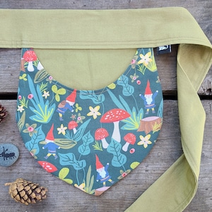 Puede incluir: Un babero de tela verde con un patrón de setas y gnomos. El babero se ata con una larga cinta de tela verde.