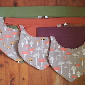 May include: Four fabric neck gaiters with a mushroom print. The gaiters are in different colors: green, orange, maroon, and gray. The mushroom print is in shades of gray, orange, and white.