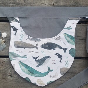 May include: A gray fabric baby carrier with a white fabric panel featuring a whale and ocean life print. The carrier has a long strap that can be tied around the waist.