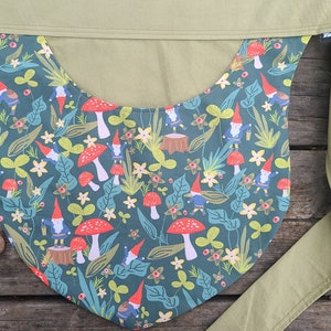 May include: A green fabric apron with a pattern of gnomes, mushrooms, and leaves. The apron has a long tie that wraps around the waist.