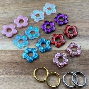 Puede incluir: Una colección de abalorios de resina con forma de flor en rosa, azul, morado y rojo con detalles de purpurina. También se muestran pendientes de aro dorados y plateados. Ideal para la creación de joyas.