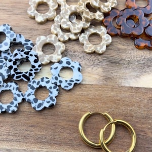Peut inclure: Diverses boucles d'oreilles en forme de fleurs aux couleurs et motifs variés. Les boucles d'oreilles comprennent des motifs tachetés noirs et blancs, beige et crème, et écaille de tortue. Une paire de créoles dorées est également visible.