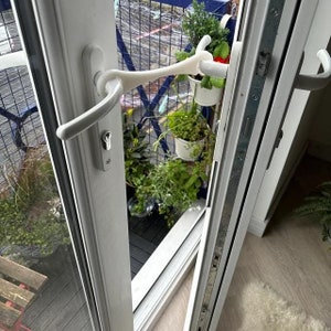Op de afbeelding: Witte deur met een witte deurknop en een witte deurstop. De deur staat open en er staan planten in potten op het balkon buiten.