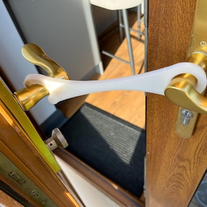 May include: A white plastic door handle opener attached to a gold door handle. The opener is designed to help people open doors without touching the handle directly.