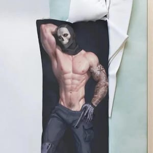 Pode incluir: Uma almofada de corpo preta com uma figura musculosa com uma máscara de caveira, roupa preta e uma tatuagem no braço. A figura está segurando uma arma. A almofada está em uma cama com roupa de cama branca e azul claro.