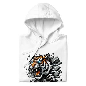 Puede incluir: Sudadera con capucha blanca con un estampado gráfico de un tigre rompiendo una pared de piedra. El tigre tiene rayas naranjas y negras y ojos azules.